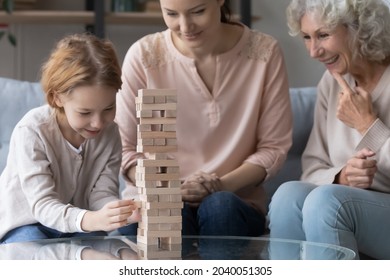 Cheerful Sincere Small Kid Girl Playing Wooden Boardgame With Young Mother And Smiling Middle Aged Mature Granny, Enjoying Carefree Leisure Weekend Pastime Together At Home, Domestic Activity Concept.