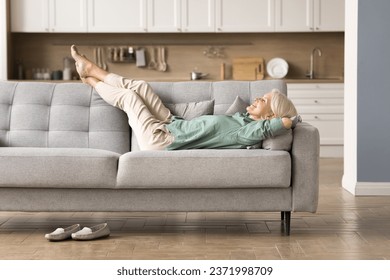 Cheerful sleepy blonde older lady lying on couch, leaning legs and feet on soft back, enjoying leisure, lazy break in modern home interior, breathing fresh cool pure air with closed eyes - Powered by Shutterstock