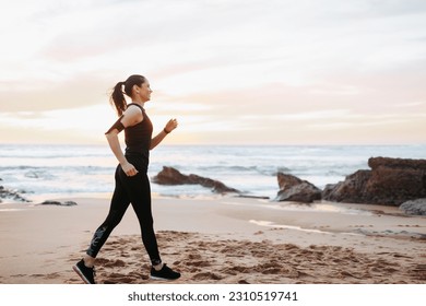 Cheerful slim adult european lady in sportswear race walking, enjoys workout in morning on sea beach, outdoor, full length. Muscles training, weight loss sport, fitness and body care - Shutterstock ID 2310519741