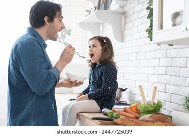 Cheerful small girl daughter and her handsome young dad are having fun while cooking in kitchen at home. Father feeding his little daughter while preparing family dinner. Happy father`s day! - Powered by Shutterstock