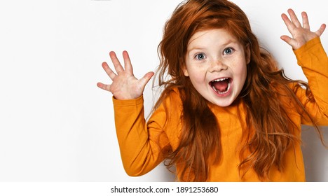 Cheerful small red haired girl with freckles in yellow comfortable longsleeve standing showing fingers and feeling excited over white background. Happy childhood, stylish look concept