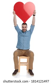 Cheerful Smart Casual Man Holding Heart Shape Above His Head And Smiling While Sitting On A Chair On White Studio Background