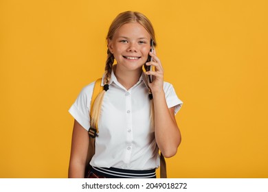 Cheerful Smart European Young Girl Student With Pigtails And Backpack Talking On Phone, Isolated On Yellow Background, Studio Shot. Technology For Learning, Back To School, Gossip And Great News