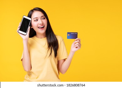 Cheerful Smile Asian Girl Holding Mobile Phone, Mockup Credit Card Transfer Internet Banking Standing On Isolated Yellow Background.Financial Young Woman Payment Online Shopping Money Transfer Concept