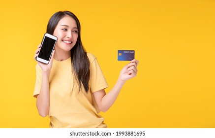 Cheerful Smile Asian Woman Holding Smartphone And Credit Card Mockup For Shopping Internet Banking Over Isolated Yellow Background. Financial Payment Online Shopping And Money Transfer Concept.