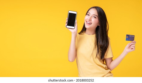 Cheerful Smile Asian Woman Showing Credit Card Mockup And Smart Phone For Internet Banking While Standing Over Isolated Yellow Background. Financial Payment Online Shopping And Money Transfer Concept