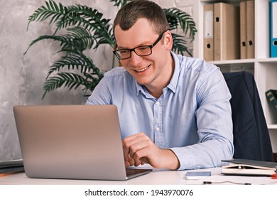 Cheerful Smiling 30 Year Old Man In Glasses And A Shirt Looks Into The Laptop Screen While Sitting In A Light Office On The Background Of A Rack With Folders. Office Work, Entrepreneurship Concept