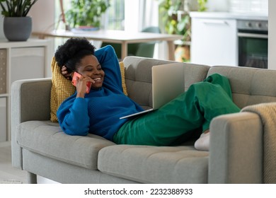 Cheerful smiling African American woman relaxing lying on sofa with laptop and talking with boyfriend on phone. Charismatic outgoing girl enjoys calling friends from college or colleagues from work - Shutterstock ID 2232388933