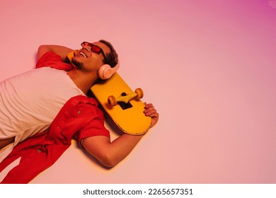 Cheerful smiling african guy in sunglasses   headphones laying and skateboard isolated over gradient neon studio background