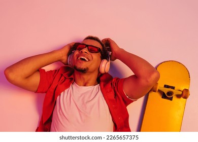 Cheerful smiling african guy in sunglasses   headphones laying and skateboard isolated over gradient neon studio background