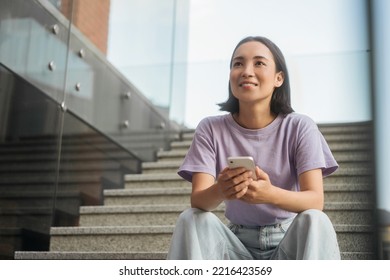 Cheerful smiling asian woman wearing purple t-shirt holding using mobile phone shopping online looking at away sitting on stairs outdoors. Technology concept  - Powered by Shutterstock