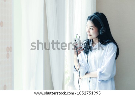 Cheerful Smiling asian young girl listening to music with headphone near window in bedroom. Beautiful,happy,  joyful, relaxing woman with earphones. Filter color effect with copyspace.