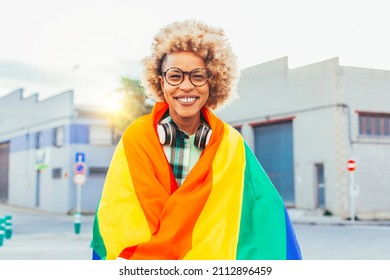 Cheerful Smiling Black Latin American Woman With Eyeglasses, Headsets, Afro Hair, Rainbow Flag Of Gay Pride On A Sunny Day Outdoors. She Fights For Sexual Freedom.