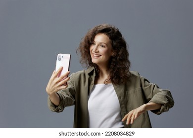 Cheerful Smiling Curly Beautiful Woman In Casual Khaki Green Shirt Doing Video Call With Friends Using Phone Posing Isolated On Over Gray Blue Background. Social Media Influencer Concept. Copy Space