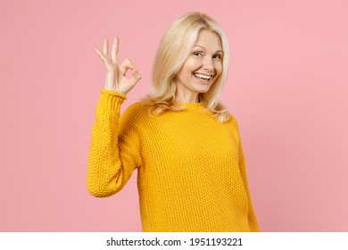 Cheerful smiling elderly gray-haired blonde woman lady 40s 50s years old in yellow casual sweater standing showing Ok gesture looking camera isolated on pastel pink color background studio portrait