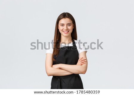 Cheerful smiling female barista in black apron cross arms chest, looking ready and confident. Young girl employee open coffee shop, greeting customers. Saleswoman running store