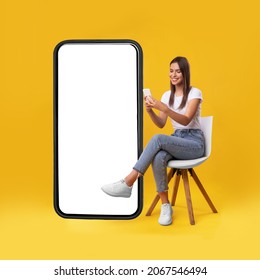 Cheerful Smiling Female Sitting On Chair Near Big Cell Phone With Empty White Screen, Using Mobile Device, Checking New Cool App On Orange Studio Background, Mock Up For Website Or Application Design