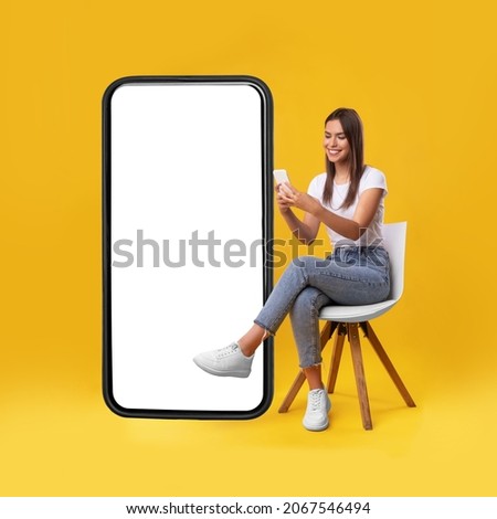 Cheerful smiling female sitting on chair near big cell phone with empty white screen, using mobile device, checking new cool app on orange studio background, mock up for website or application design