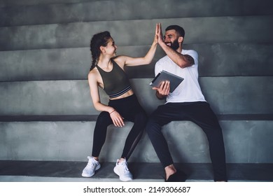 Cheerful Smiling Friends In Activewear Sitting On Stairs And Giving High Five To Each Other While Resting After Training In Gym Together