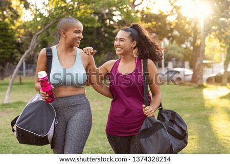 Cheerful smiling friends in sportswear holding gym bag and bottle in park. Multiethnic women going to park for fitness workout. Two curvy girls walking after exercise session outdoor at sunset.