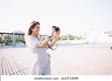 Cheerful smiling lady with long hair in light dress with polka dots taking photo with black camera standing on wooden floor in street in summer  - Shutterstock ID 1613714098