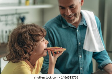 Cheerful And Smiling Little Boy Eating Toast Bread With Jam With Happy Mature Father