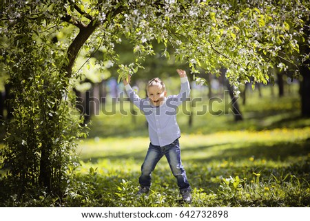 cheerful smiling little boy jump under beautiful blooming apple or cherry tree at spring time. Funny kid in blue shirt and jeans has fun in nature. Cute blonde child in spring garden sun back lighting