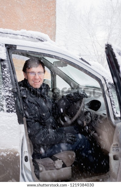 Cheerful smiling\
man in black jacket is sitting in the car driving during heavy\
snowfall and looking at\
camera.