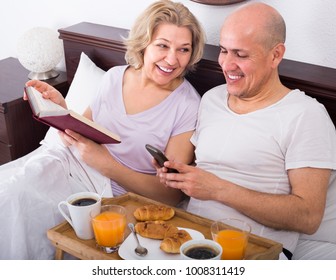 Cheerful smiling mature couple reading book while having breakfast in bed. Focus on woman
 - Powered by Shutterstock