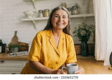 Cheerful smiling mature female in good mood drinking tea on kitchen. Joyful senior lady in yellow dress posing for picture indoors. Rest, morning, happiness, growing old concept