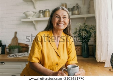 Cheerful smiling mature female in good mood drinking tea on kitchen. Joyful senior lady in yellow dress posing for picture indoors. Rest, morning, happiness, growing old concept