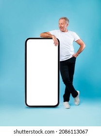 Cheerful smiling mature man looking at giant big cell phone, leaning on empty white screen template panel, standing on blue studio wall. Mock up for app or website, free copy space, full body length