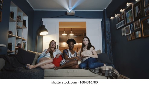 Cheerful smiling multiethnic female friends watch film together at home, laugh and talk eating snacks slow motion.