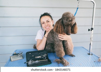 Cheerful Smiling Professional Female Groomer Sitting Near Cute Brown Poodle Dog After Haircutting. Animal Hair Cut In Pet Care Salon. Small Business Owner Concept. 