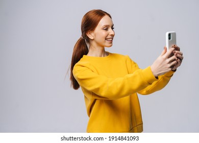 Cheerful Smiling Redhead Young Woman Having Video Call With Lover Holding Smart Phone In Hand Shooting Selfie On Front Camera Isolated On White Background.