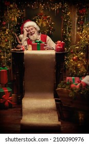 Cheerful smiling Santa Claus thinks about a list of good and naughty children sitting in festively decorated room with a gift box and a long magic scroll in front of him. Christmas fairy tale. 