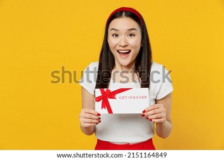 Cheerful smiling shocked young girl woman of Asian ethnicity 20s years old wears white t-shirt hold gift certificate coupon voucher card for store isolated on plain yellow background studio portrait