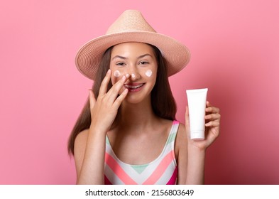 Cheerful smiling tanned teen girl put sunscreen on her face and holds a white blank cream tube mock up in hand, wear swimsuit and straw hat isolated on pink background sun protection skin care concept