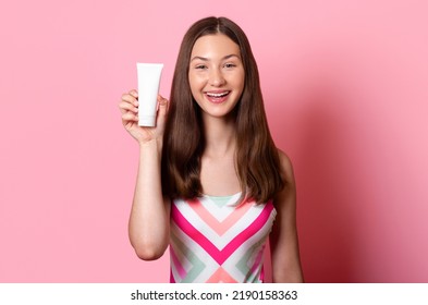 Cheerful smiling tanned teen girl hold in hand white blank cream tube mock up sunscreen, wearing colorful one piece swimsuit isolated on pink background