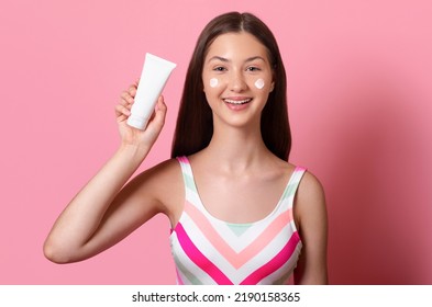 Cheerful smiling tanned teen girl hold in hand white blank cream tube mock up sunscreen, wearing colorful one piece swimsuit isolated on pink background