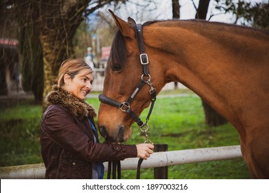Cheerful and smiling woman dressed in a brown jacket looking at her horse at the ranch