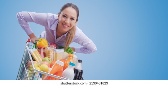 Cheerful smiling woman leaning on a full shopping cart, grocery shopping concept - Powered by Shutterstock