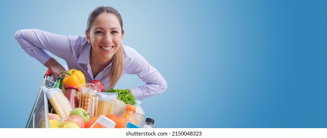 Cheerful smiling woman leaning on a full shopping cart, grocery shopping concept