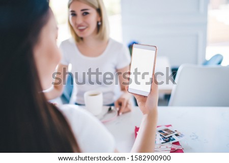 Cheerful smiling women in white t shirts sitting at table texting on mobile phone drinking cups of coffee in modern white cafeteria 