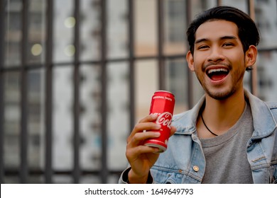 Cheerful Smiling Young Asian Hipster Man With Coca Cola Drink In Hand. Man Holding And Drink Coke Can With Happy  Face At Sunset. BANGKOK,THAILAND - FEBRUARY 2 2020.