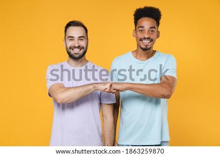 Cheerful smiling young friends european african american men 20s wearing casual violet blue t-shirts giving fists bump looking camera isolated on bright yellow colour wall background studio portrait