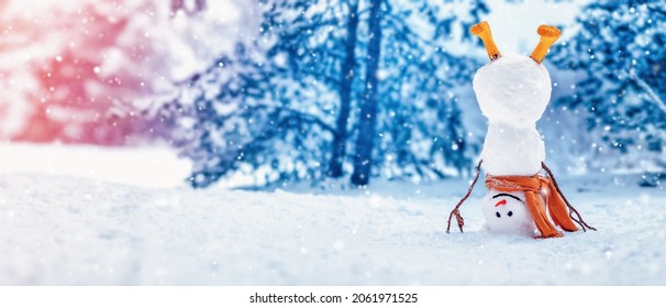 Cheerful snowman with orange scarf and in yellow boots stands upside down in winter forest