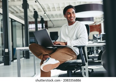 Cheerful software developer smiling in an office. Happy young businessman looking away while working on a laptop in a modern workplace. Creative businessman working on a new project.