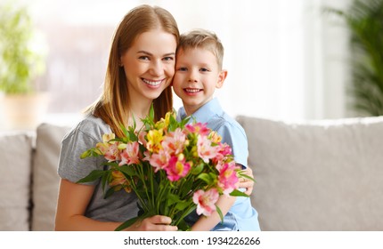 Cheerful Son Embracing Mother Sitting On Sofa With Bouquet Of Alstroemeria Flowers On Mothers Day