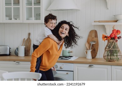 Cheerful son at kitchen riding  on moms back laughs happy spend time together. Curly Spanish woman I orange sweater entertains little boy home. Playful Italian kid hugs babysitter. Happy family. - Powered by Shutterstock